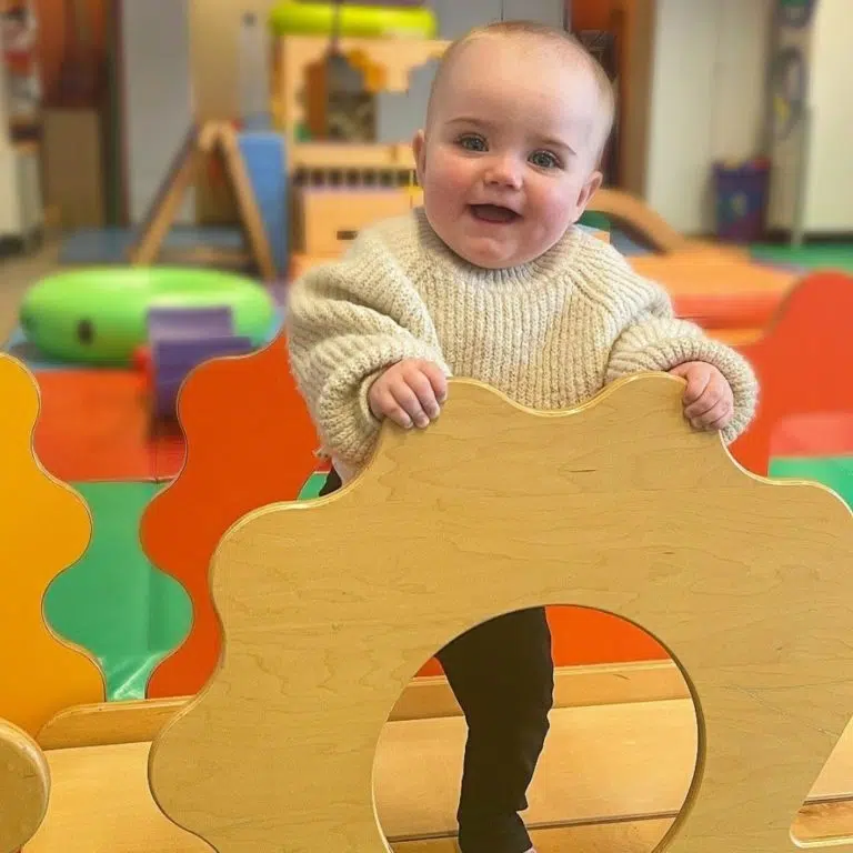 A 12 month old baby standing up holding on to a climbing frame, smiling at the camera